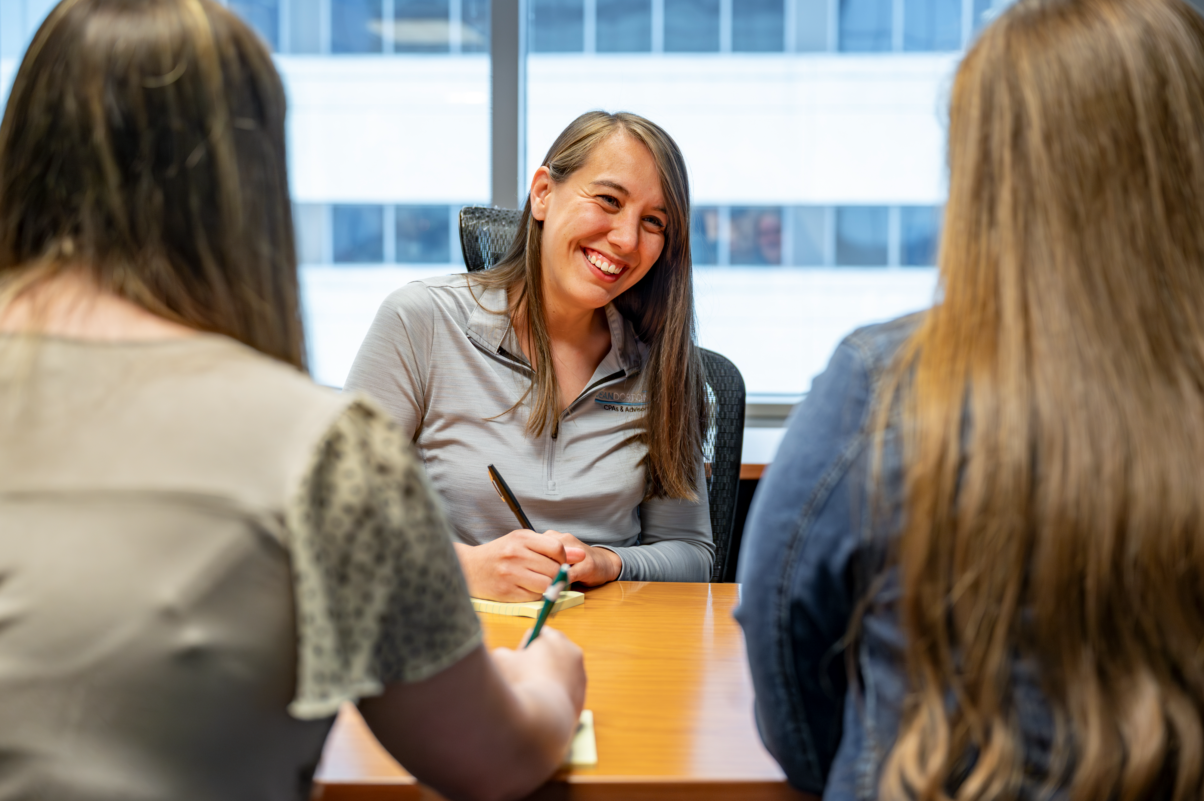 A smiling young woman sits at a table and speaks to two other women sitting across from her as she does paperwork.