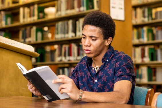 A young man sits at a table in a library and reads a book.