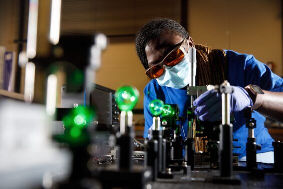 A woman in a mask an protective glasses carefully works with small manufacturing equipment.