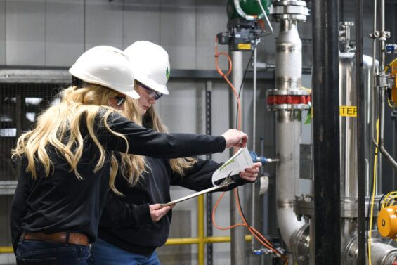 Two women in hard hats stand in a warehouse and look at a clipboard together.