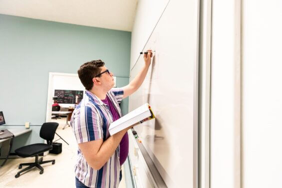 A man is focused as he writes on a whiteboard while holding a book in his other hand.